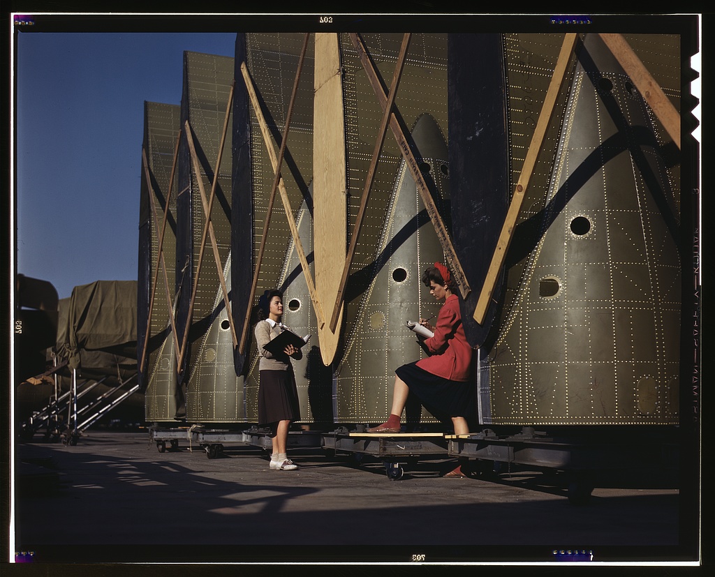 Two inspectors with clipboards checking the inner wings of cargo planes lined up in a factory yard.